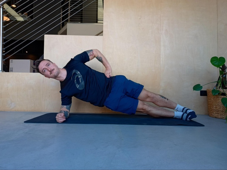 Man On Yoga Mat Showing Someone How To Do The Side Plank Exercise